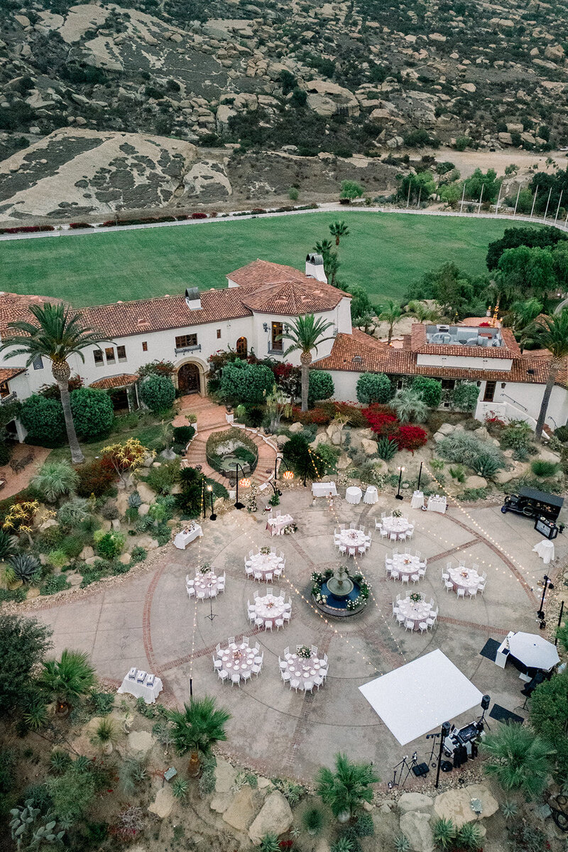 Aerial Drone view of an editorial-style wedding ceremony at Hummingbird Nest Ranch in Simi Valley, CA captured by a film wedding photographer. Guests are seated in a circular courtyard surrounded by palm trees and bougainvillea.