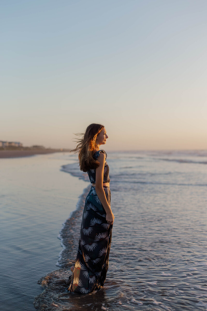 Beach girl about to turn 16 stands in the water in a blue dress looking at the sunrise over the water
