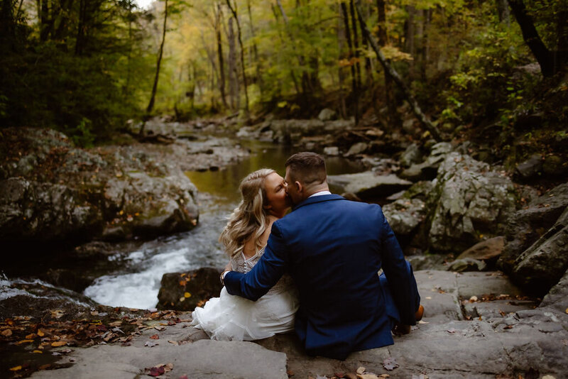 Cades Cove in the Smoky Mountains with bride nad groom sitting on top of a waterfall together with the grooms arm around the bride and kissing her with a river in front of the couple captured by gatlinburg photographers