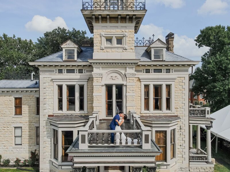 A couple embraces on the balcony of an ornate stone mansion with a distinctive tower, under a clear sky at a scenic Iowa wedding.