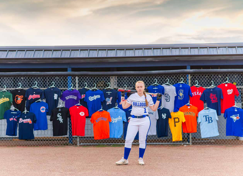 high school senior girl in softball uniform for ionia high school, standing in front of  a fence covered in jerseys