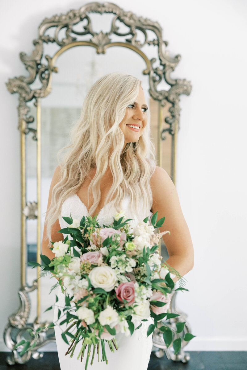 Bride stands in front of mirror and smiles holding bouquet at wedding venue in Mechanicsburg, PA