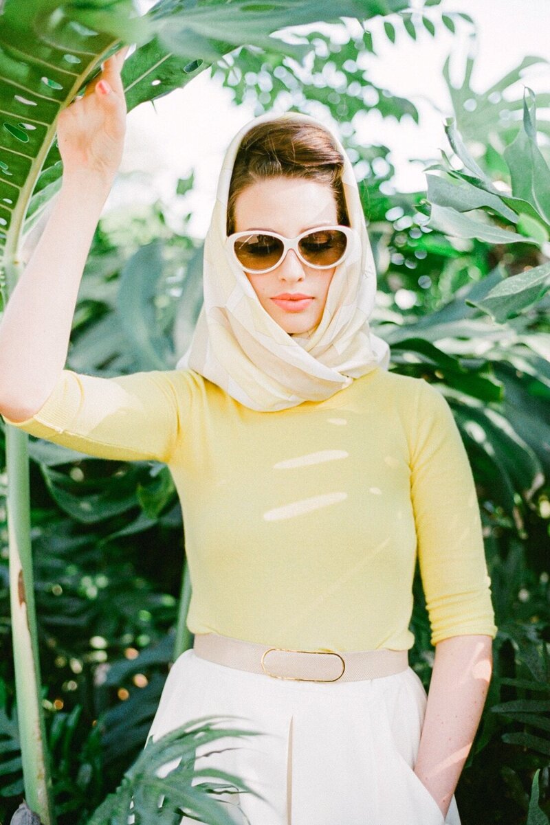 styled 50s vintage portrait of a woman  wearing a headscarf, cat-eye sunglasses, and posing under huge monstera leaves on Maui