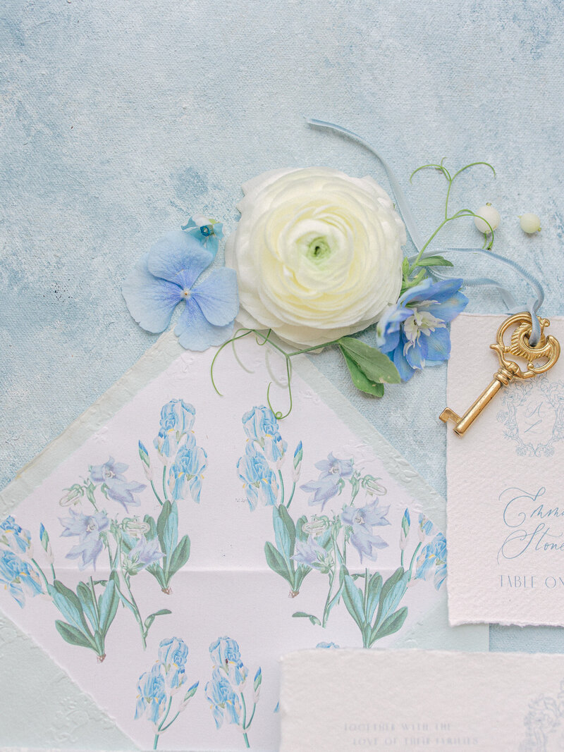 Close up of the inside of a floral wedding invitation envelope with blue and white flowers and a gold key beside it on a light blue textured backround