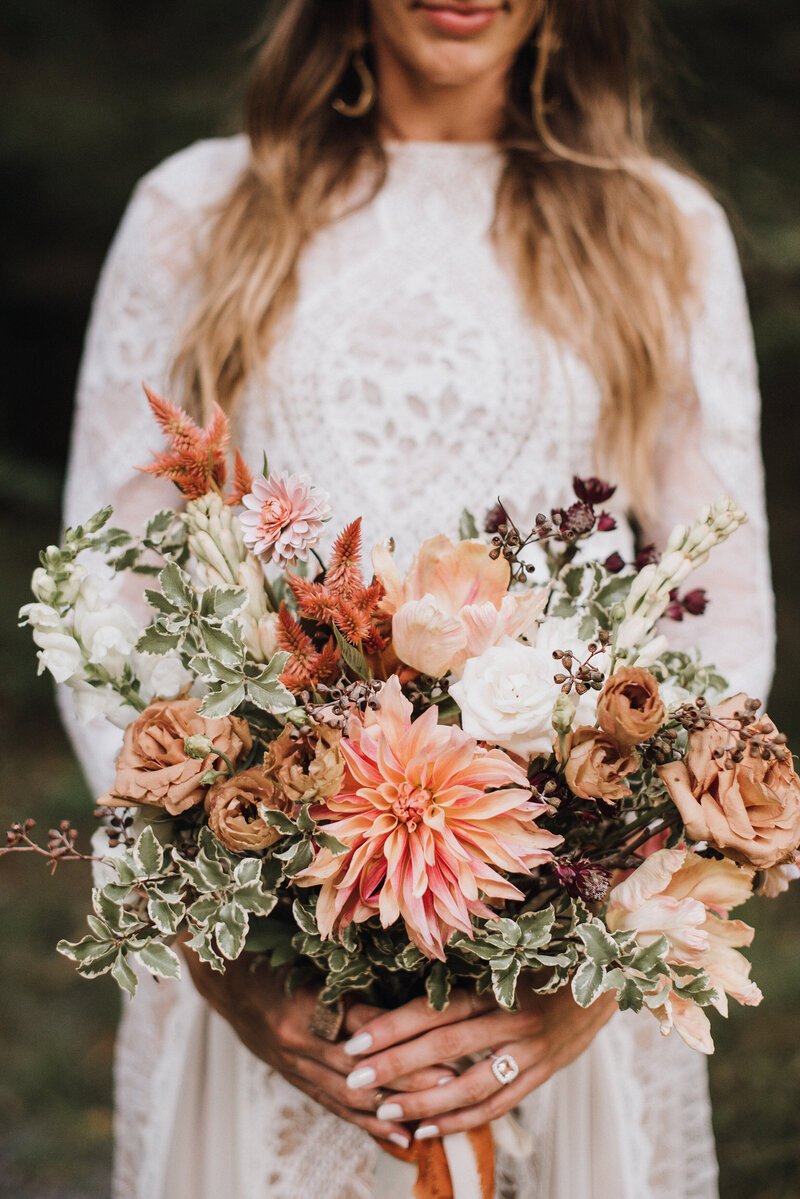 A bride in a lace wedding dress carries a bouquet of peach dahlias copper celosia tuberose brown lisianthuus toffee roses and peach tulips tied with silk ribbon