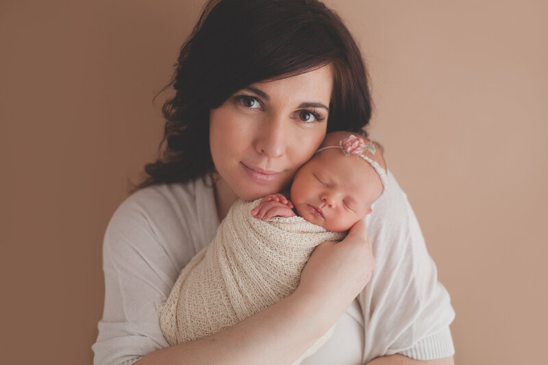 beautiful mother holding beautiful baby girl while looking into camera with tan background