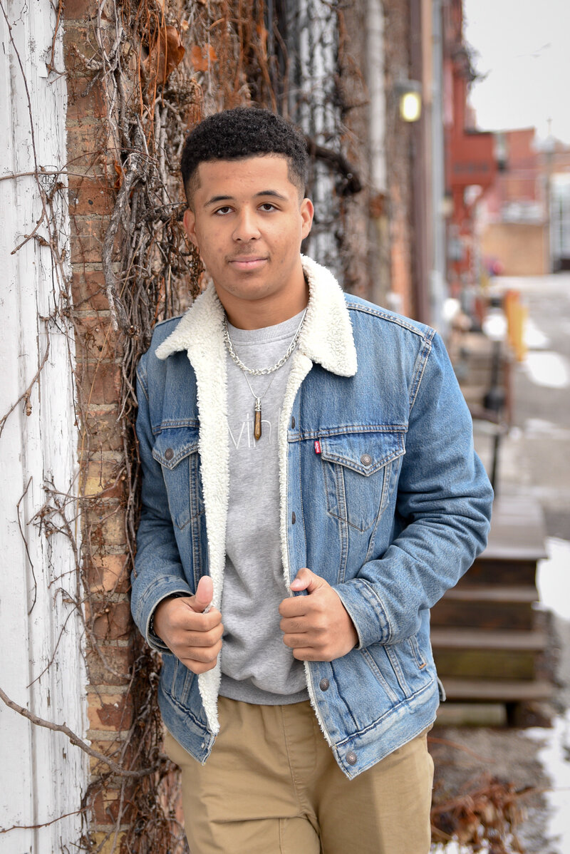 Teenage boy poses for senior portrait in denim jacket against brick wall
