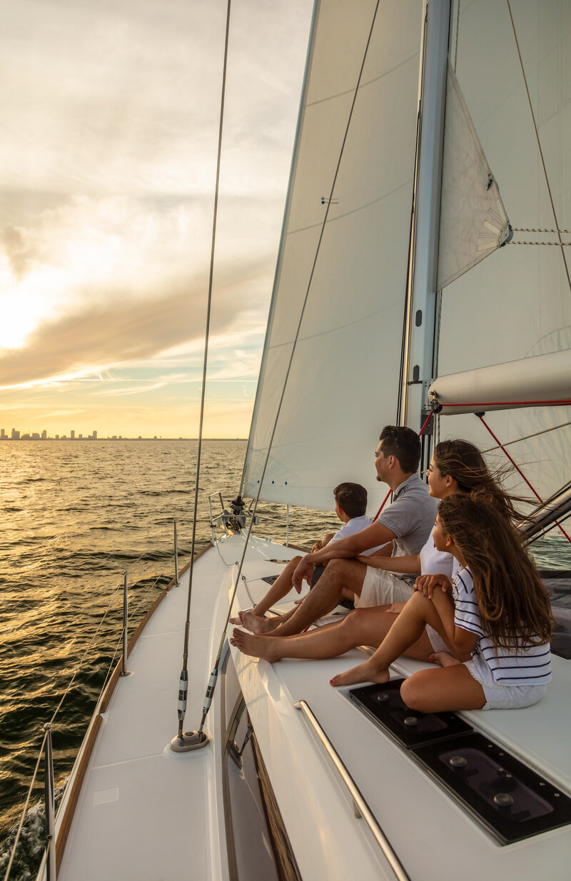 A family of four sits together at the bow of a sailboat, gazing at the sunset over the ocean. The city skyline is visible in the distance, and the sails are fully extended as the boat glides through the water.