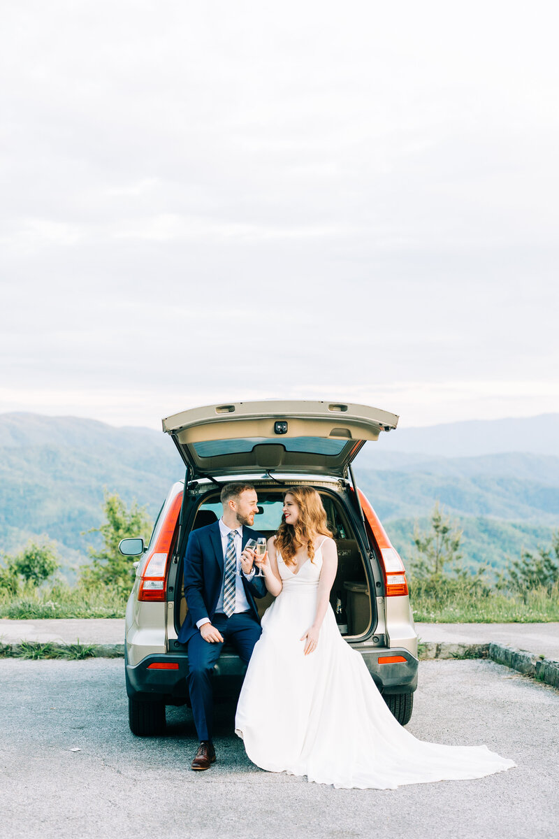 Bride and Groom sit in the back of their car overlooking the mountain,  smiling at eachother