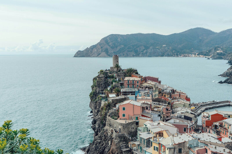 Scenic view of a coastal town on a rocky cliff overlooking the sea. Colorful buildings are clustered along the cliffside, with a circular tower near the edge. Hills and a cloudy sky are visible in the background.