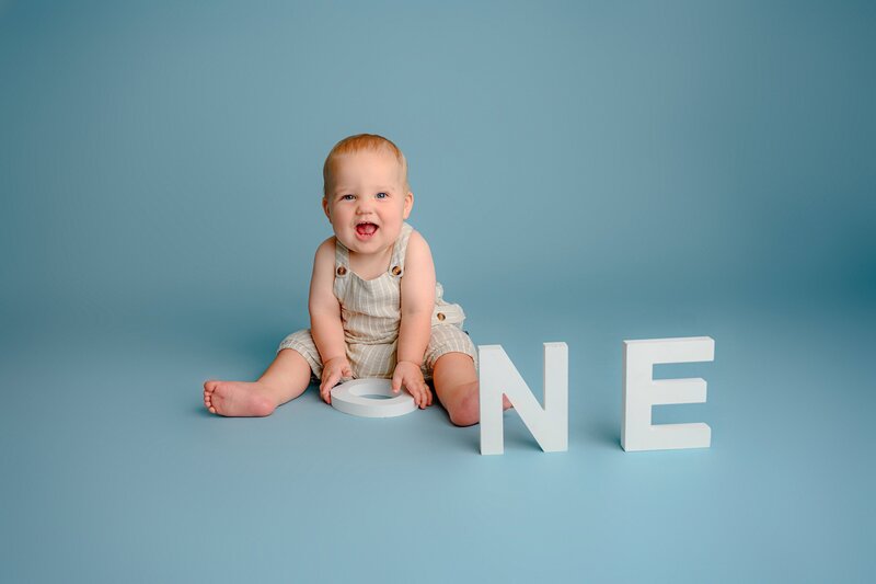 little boy sits in brown overalls playing with  ONE letters on a blue studio background