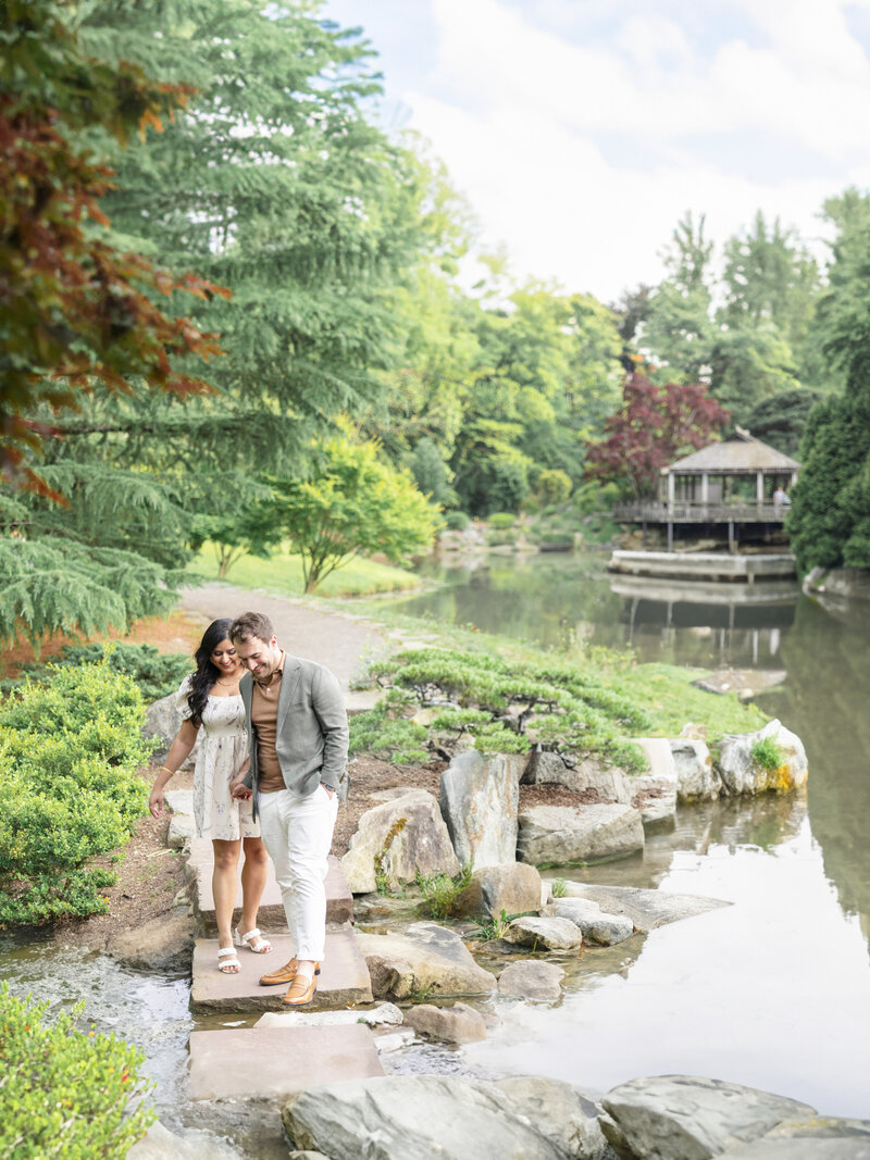 A couple walks arm in arm over stepping stones next to a serene pond in a lush garden. Trees and greenery surround them, with a wooden gazebo visible in the background, reflecting in the water. The scene conveys a peaceful and romantic atmosphere.