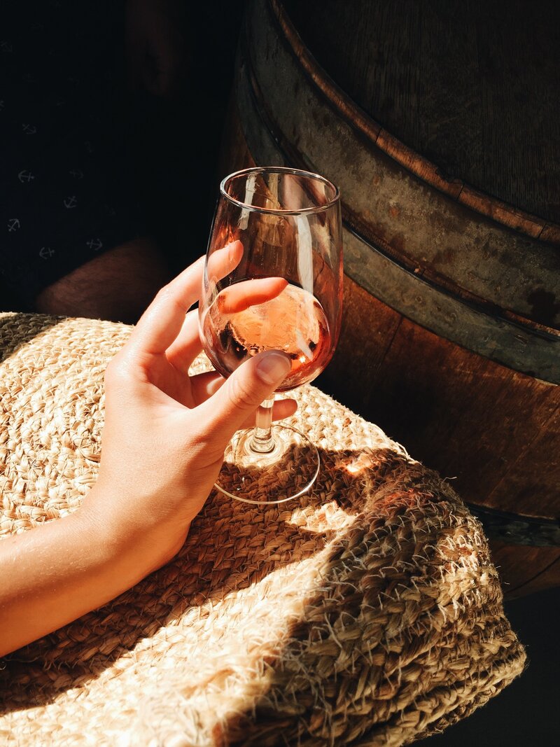 A woman's hand holds a glass of rose in a sunspot on top of a woven mat.