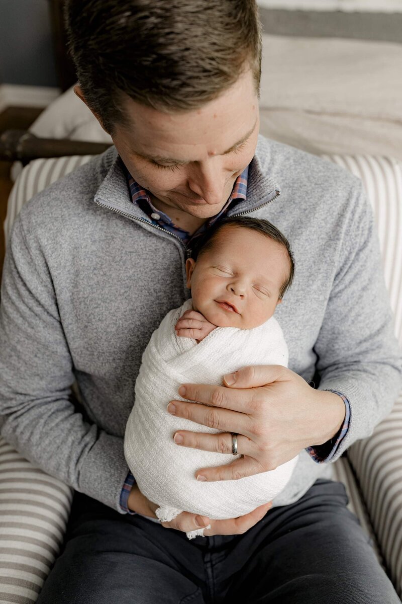 Father looks down towards his newborn son who is wrapped in White in their Charlotte Area home during their lifestyle newborn session