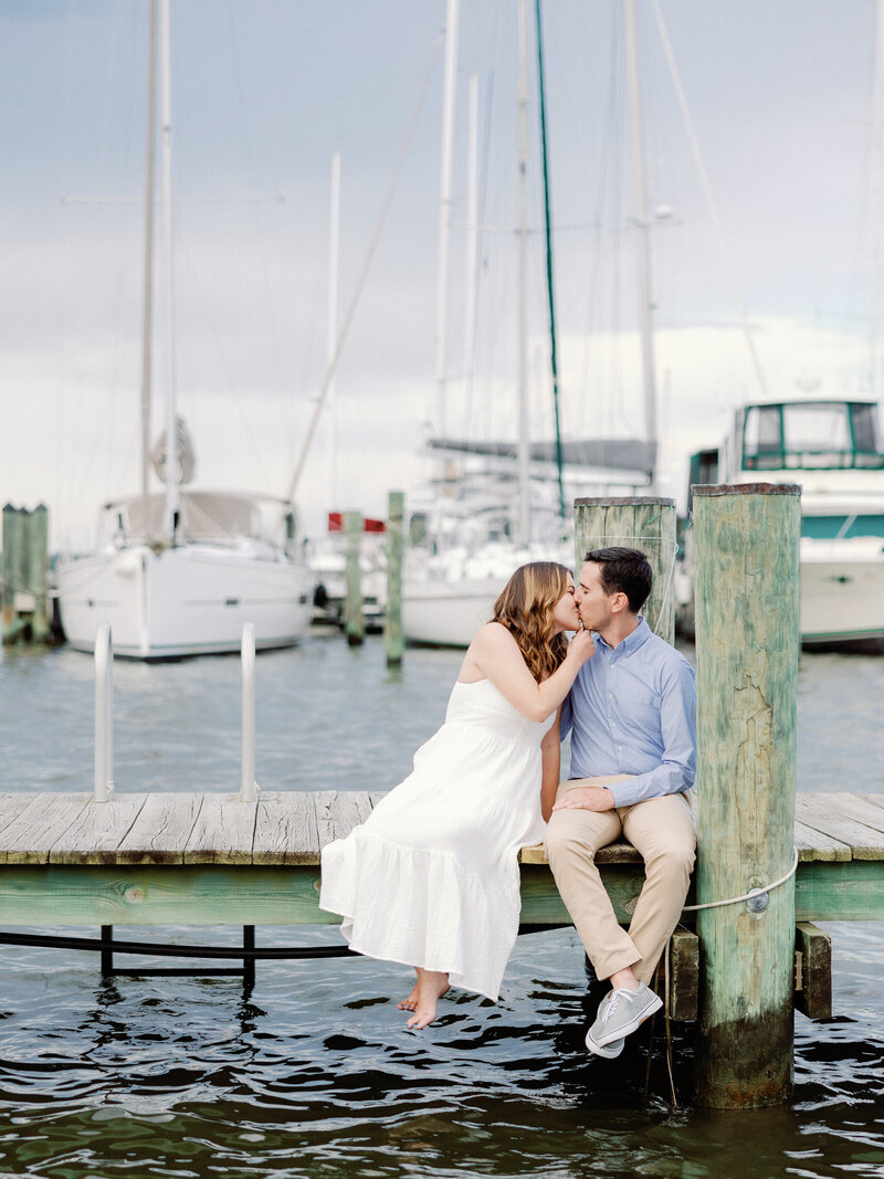 A couple shares a kiss as they sit on a dock at the Annapolis maritime museum and park with sailboats and water in the background