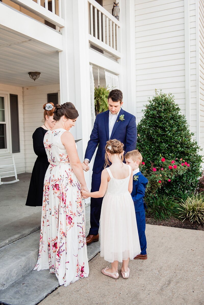 The bride and groom hold hands with the flower girl and ring bearer as they pray during their wedding ceremony. The officiant is wearing a black dress with a silver flower in her hair. The bride is wearing a sleeveless floor length white dress with pink flowers. The flower girl is wearing a long white sun dress. The groom and ring bearer are wearing matching cobalt blue suits.  They  are standing on the front porch of a large white mansion.