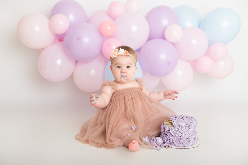Baby eating a purple rosette cake with a pink purple and blue balloon garland