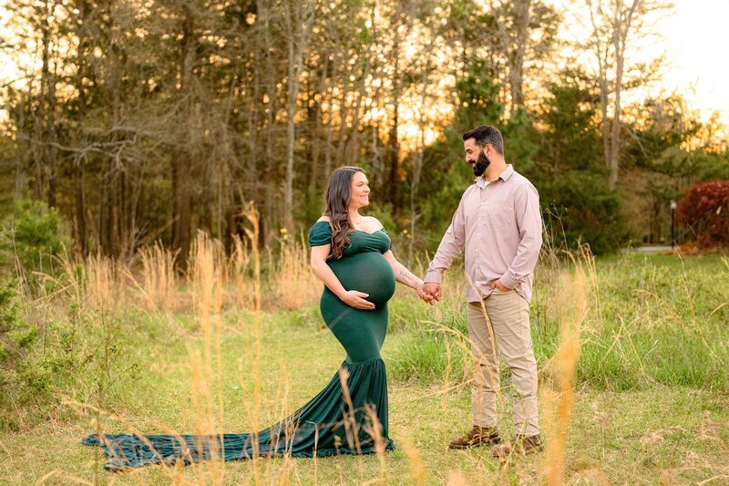 Charlotte couple stands hand in hand looking at each other in a field mom is dressed in a hunter green maternity dress with one hand on her belly dad is dressed in a button down and pants with his hand in his pocket