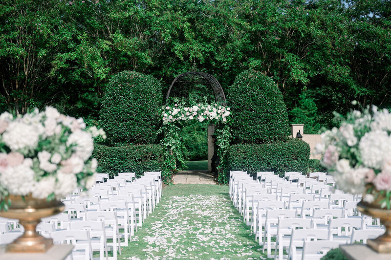 bride and groom wedding ceremony in lush garden with white chairs and flower arch