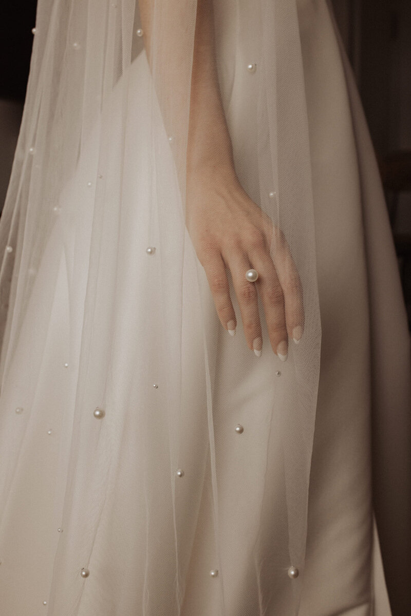 Close-up photo of a bride's hand on her gown, pearls on veil in sharp detail.