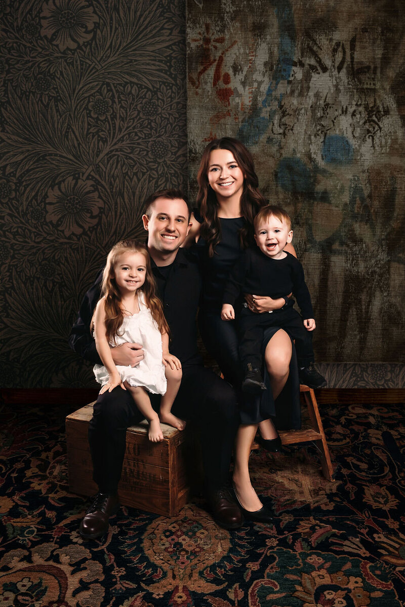 A family of four poses in a studio in Bemidji, Minnesota; two adults and two young children sit on wooden boxes and ladders against patterned wallpaper and a carpeted floor, captured by a local photographer.