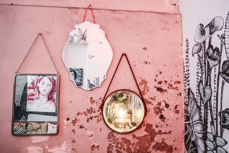 A framed sketch and photo and two mirrors reflecting artwork and plantlife hang from a richly textured pink and red wall