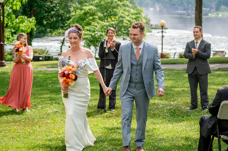 Bride and groom walking hand in hand on green lawn surrounded by family near the water – photographed by wedding photographer Niko Coric – Lumen Clarity Media