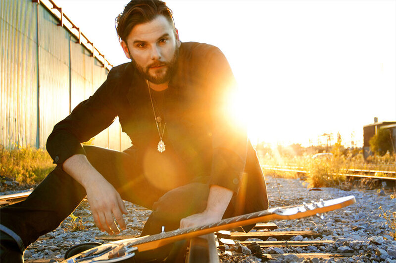 Musician portrait Brad James sitting on gravel between train tracks with guitar sun set burst behind him
