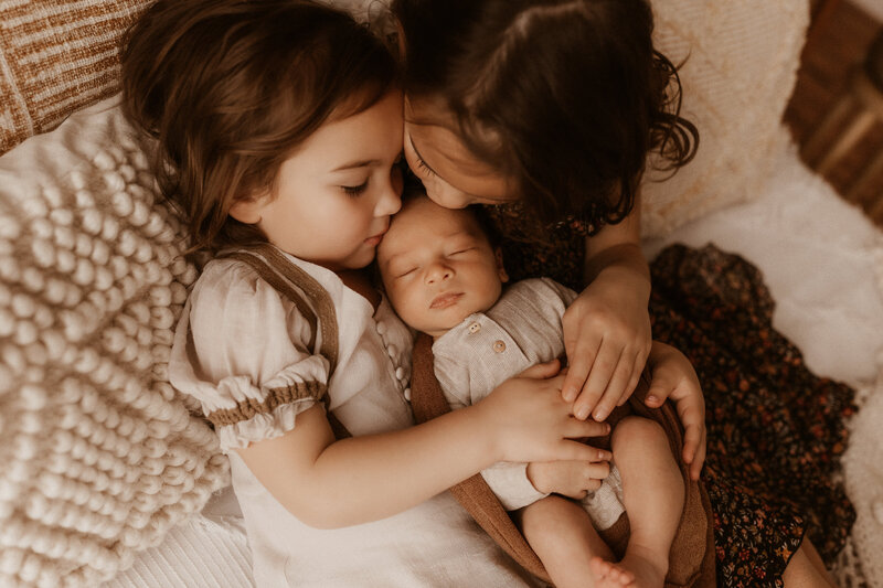 Two holder sisters snuggle their newborn baby brother in bed.