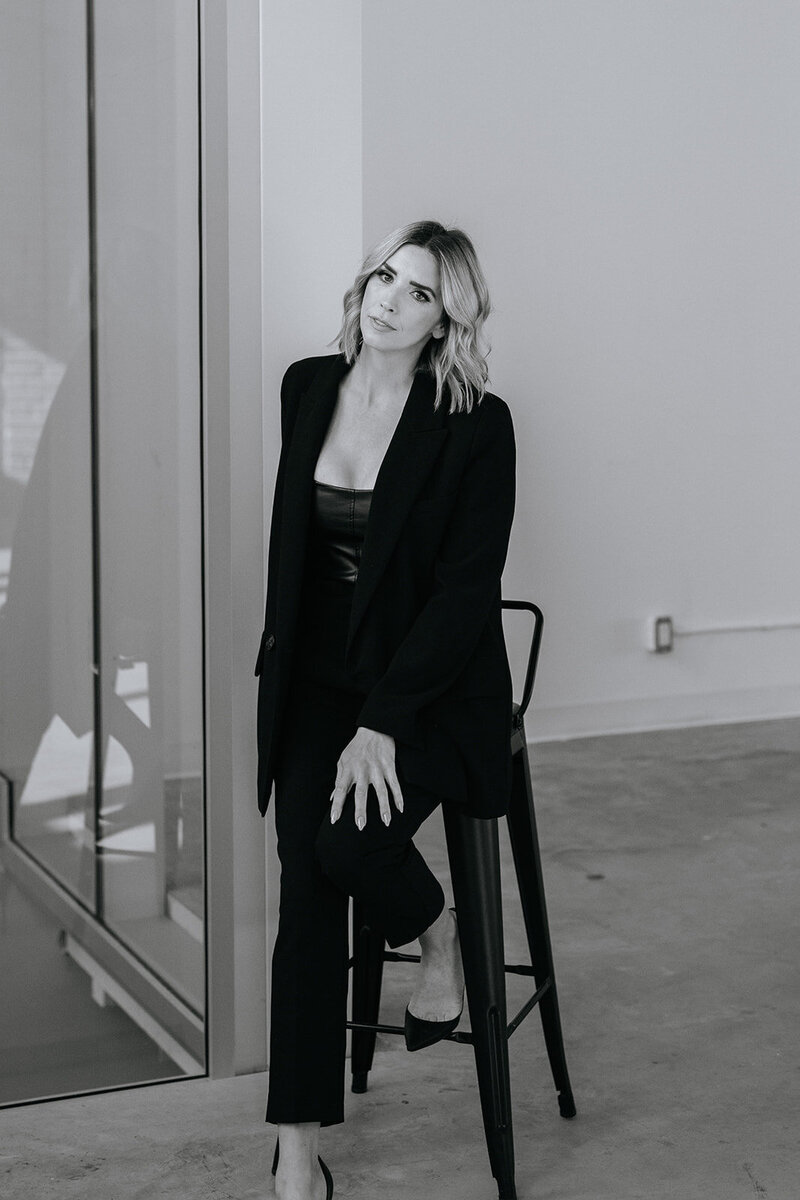 Black and white portrait of woman sitting on a stool in a black suit.