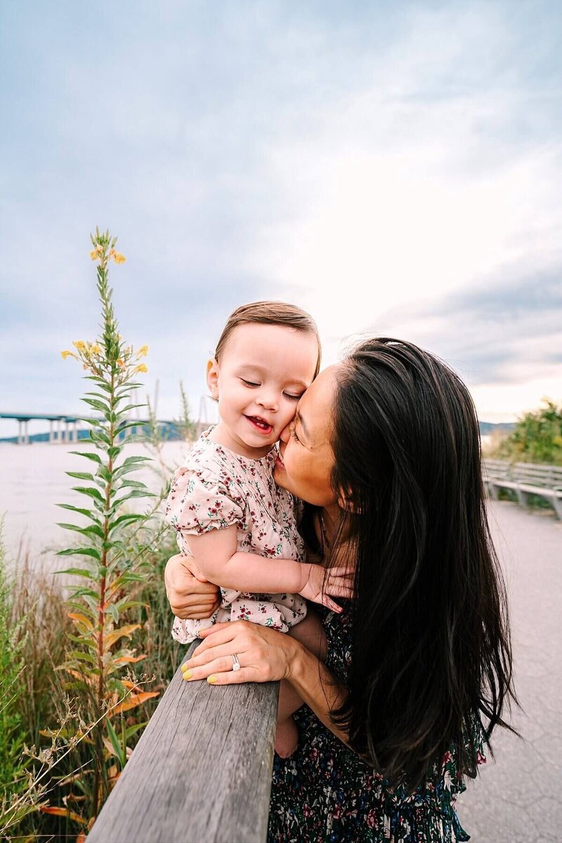A woman with long, dark hair kisses a smiling toddler on the cheek by a wooden railing, with a river and a bridge in the background under a bright sky.