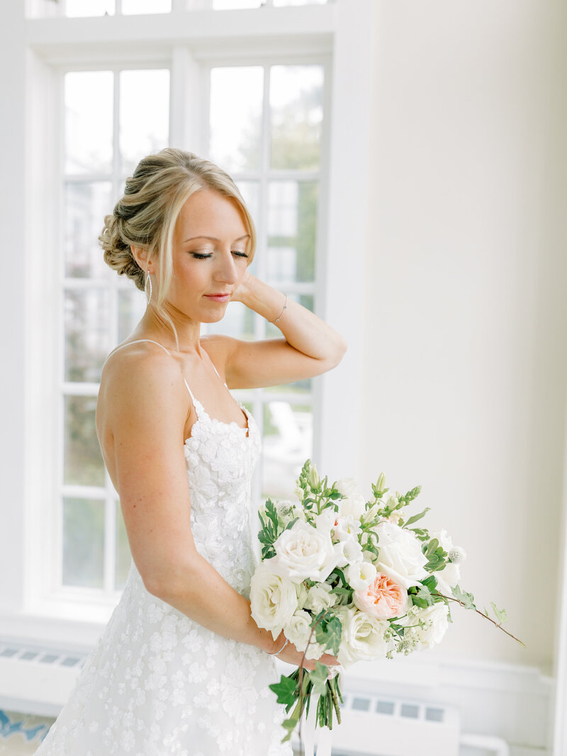 Portrait of bride standing beside a window holding her white and green bouquet and touching her hair