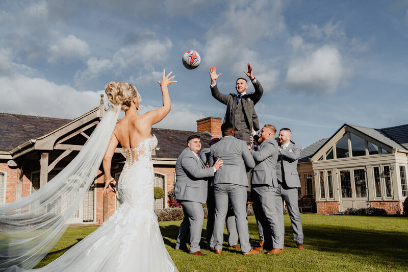  A playful wedding moment where the bride throws a rugby ball and a group of groomsmen lift the groom to catch it, captured at a destination wedding in Europe by a UK wedding photographer and videographer.
