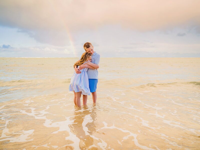 Brother and sister smile and embrace under a rainbow in the ocean on Oahu during Love + Water Photography shoot at sunset