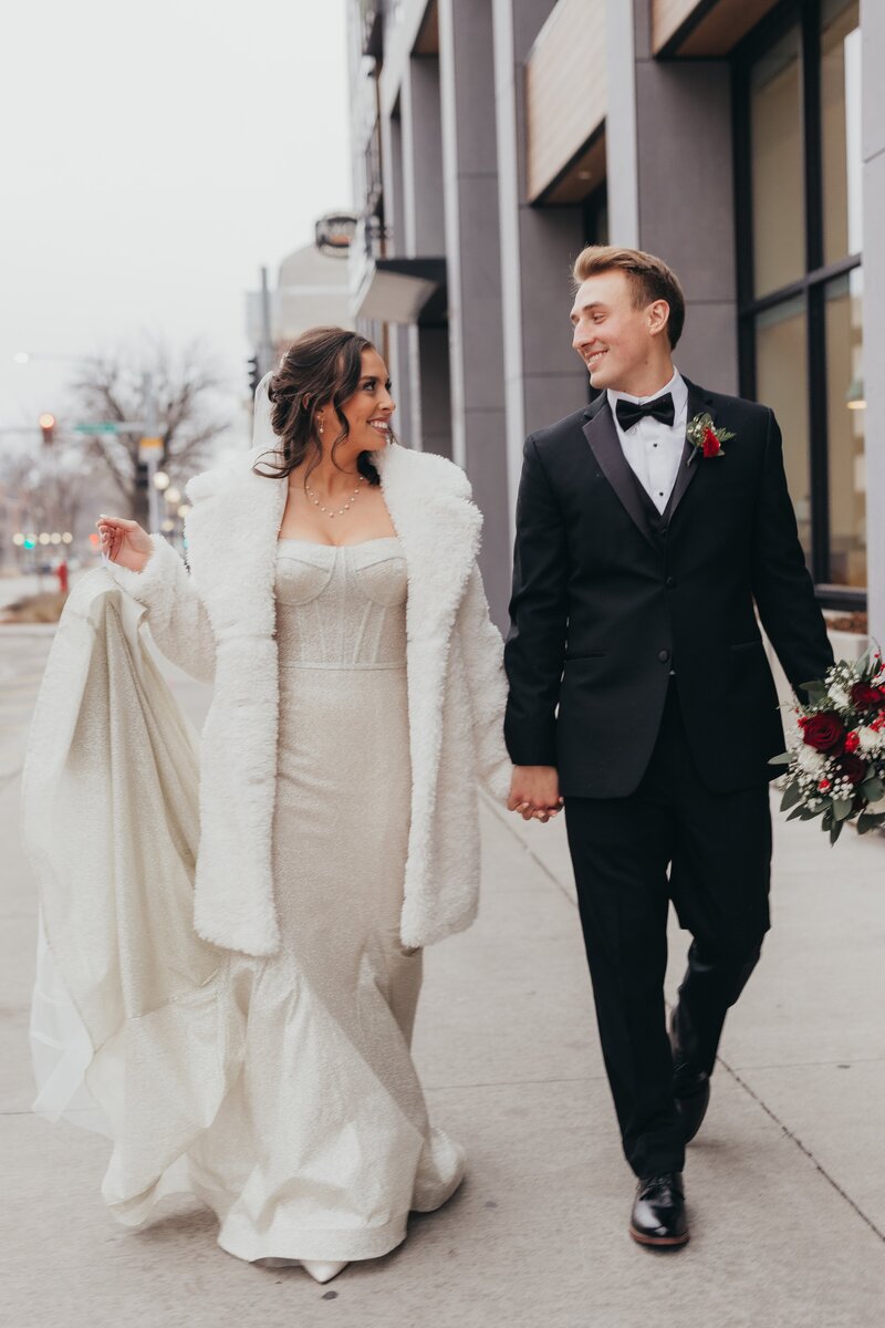 A bride in a white dress and fur stole and a groom in a black tuxedo smiling and holding hands while walking down a city street at an Iowa wedding.