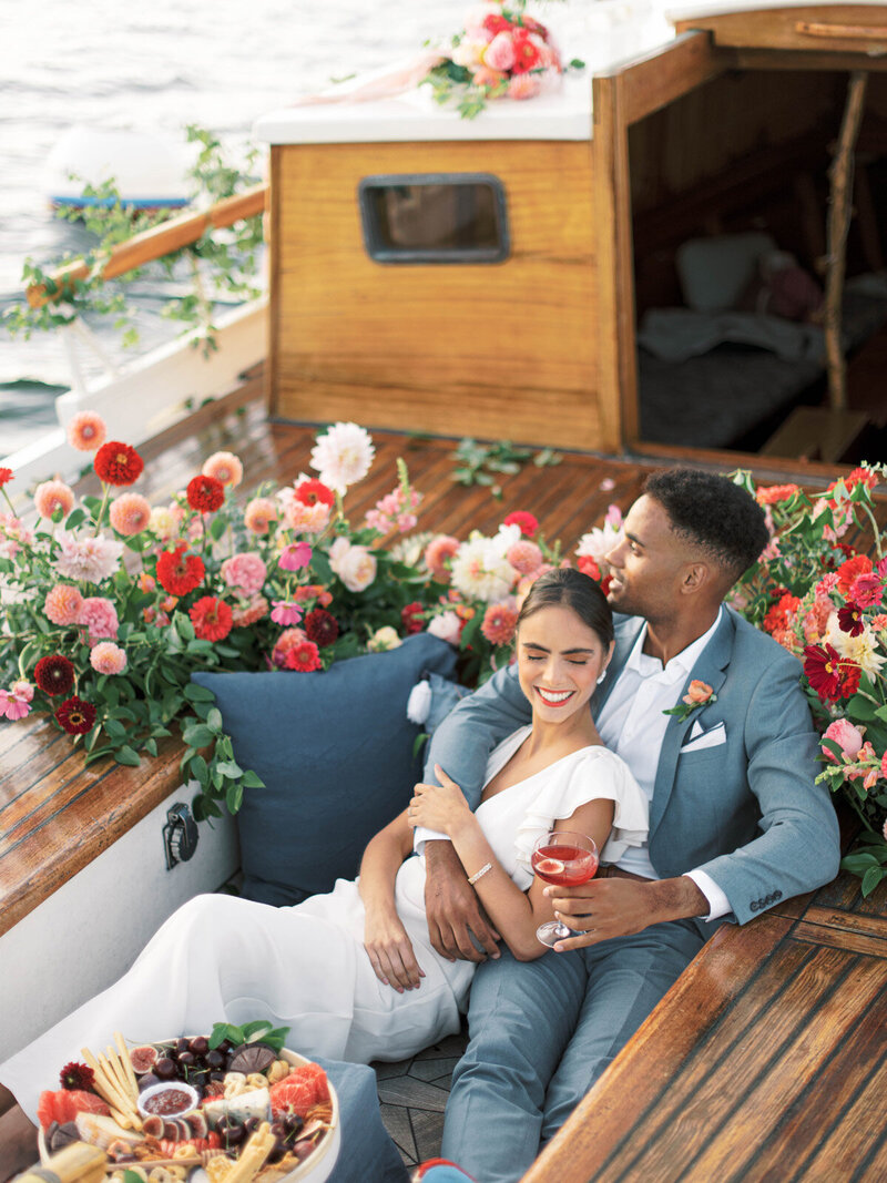 A bride and groom sit on a yacht cuddling, flowers surround them