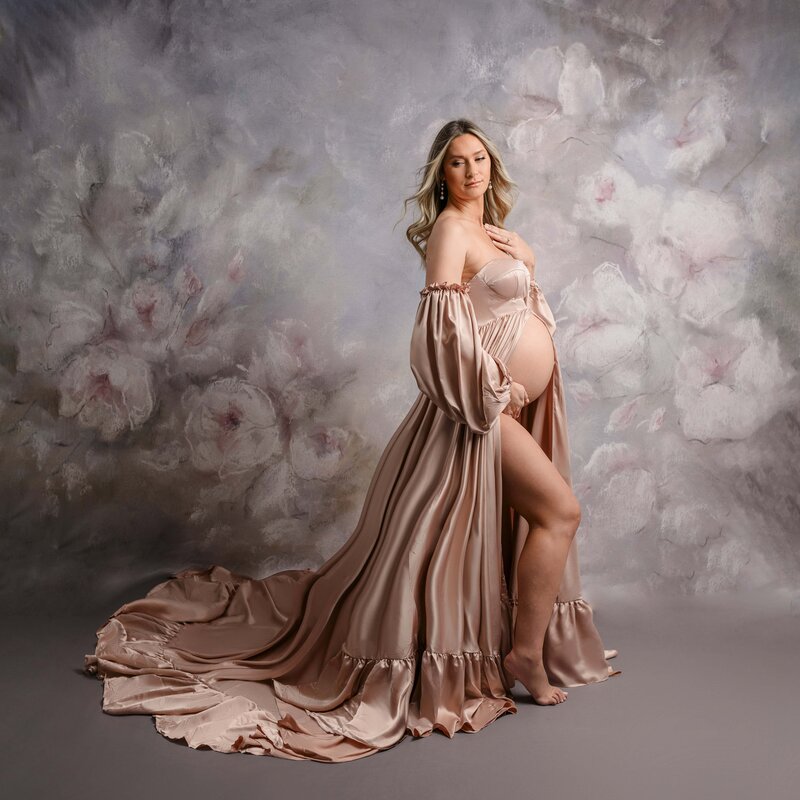 A pregnant woman stands against a floral background, wearing a flowing, strapless pink maternity  gown that drapes elegantly to the floor.