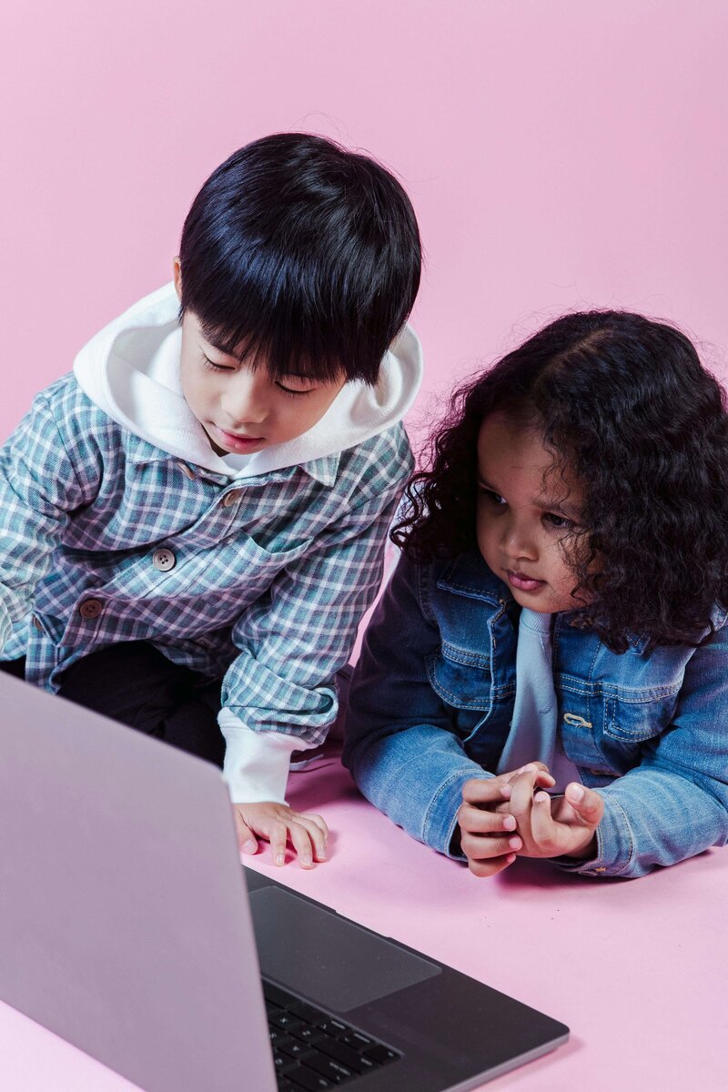 Two young children focused on a laptop screen while sitting on a pink background, wearing casual clothes.