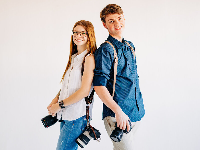 This image features Kylie and Daniel, owners of KD Captures, a wedding photography and videography business in San Antonio, TX. The image features a man sitting on a stool while holding a gimbal with a camera attached, and a woman leaning on him while holding her own camera. The man is wearing a grey shirt and khaki pants, and the woman is wearing a black crop top and jeans.