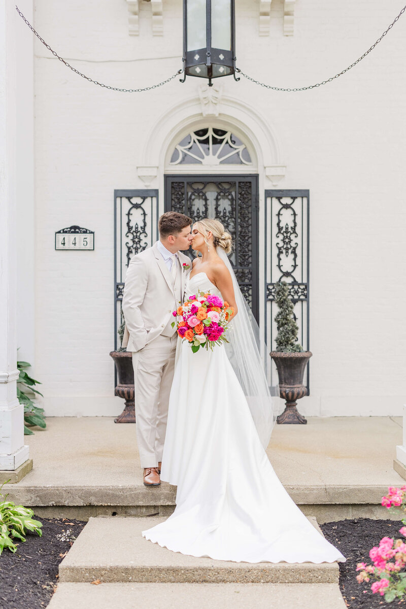 A bride and groom kiss while standing on a the stairs of a big white house. She is holding a bright pink bouquet and he is wearing a tan suit.