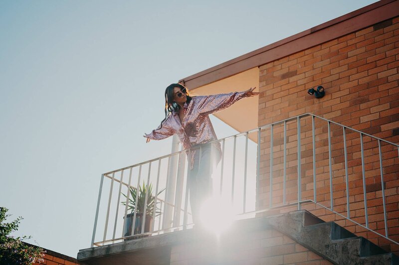 Creative branding photo of Sharni Tolhurst on a balcony wearing a sequin top, reaching into the sunlight.