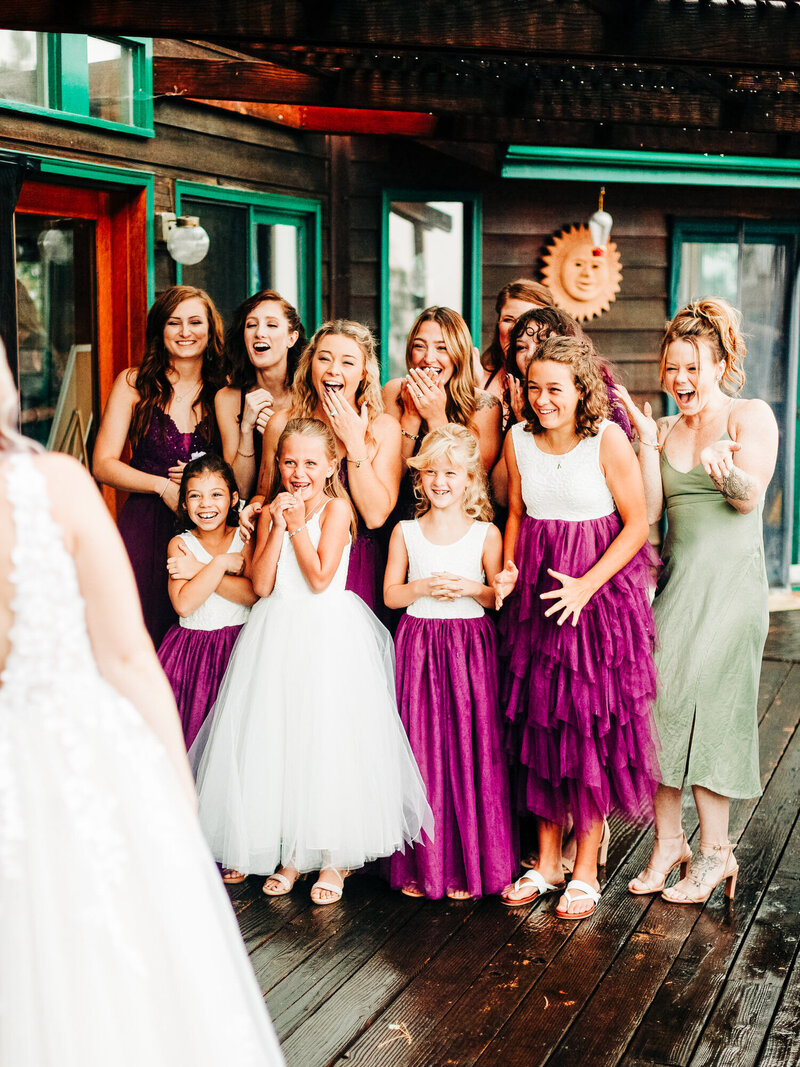 A bridal party reacts to seeing the bride for the first time. The flower girls are wearing dresses with purple skirts and white tops, and the bridesmaids are wearing purple dresses. The image was taken at the ceremony venue, the bride's family's backyard. The bridesmaids and flower girls are all happy and suprised. The image was taken by Wedding Photographer KD Captures, based out of San Antonio, TX.