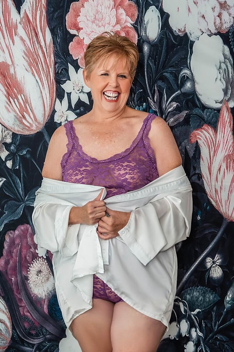 Older woman smiles as she stands in front of a floral backdrop during a boudoir session.
