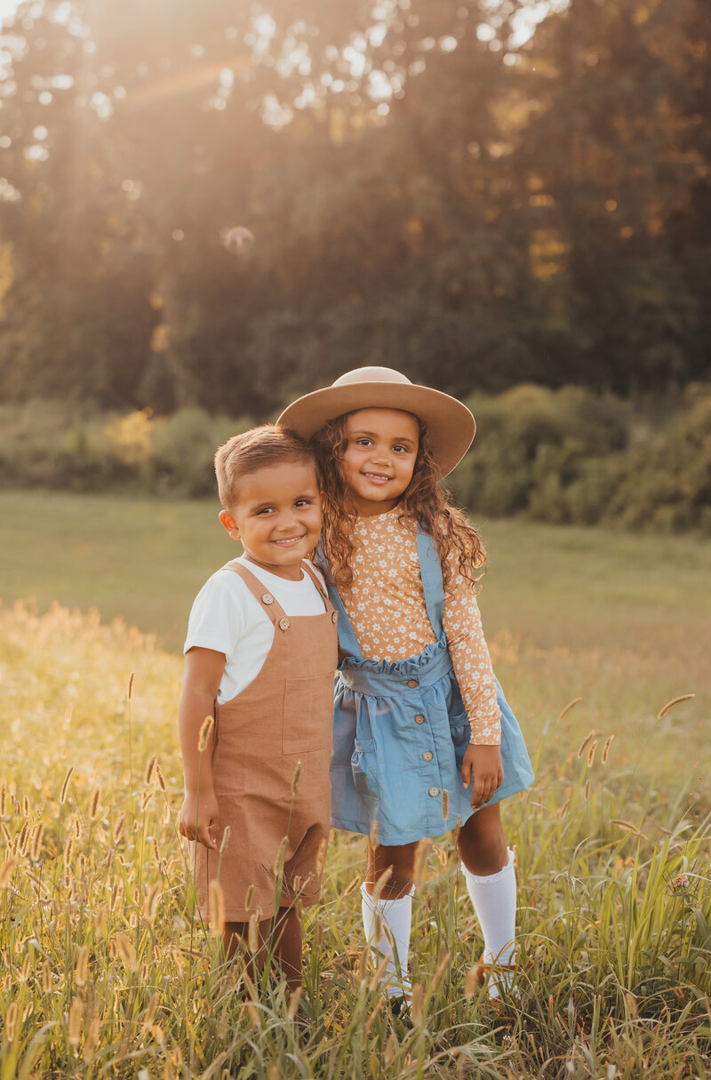 Family photo of little girl and boy standing together in field of grass during golden hour