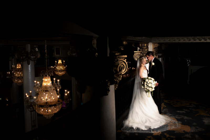 Bride and groom gaze at each other from a balcony with dramatic chandeliers