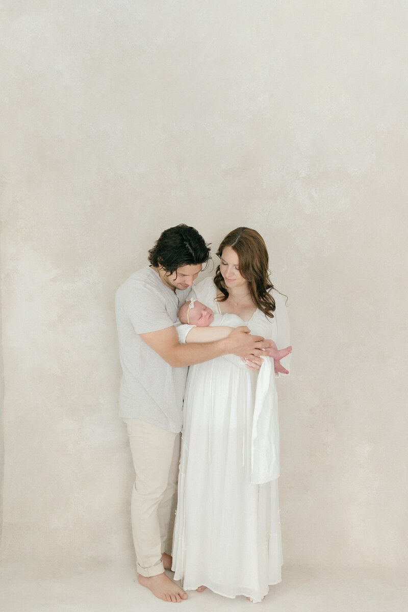 A mom and dad holding their newborn baby while gazing down at her in all white studio by NJ photographer