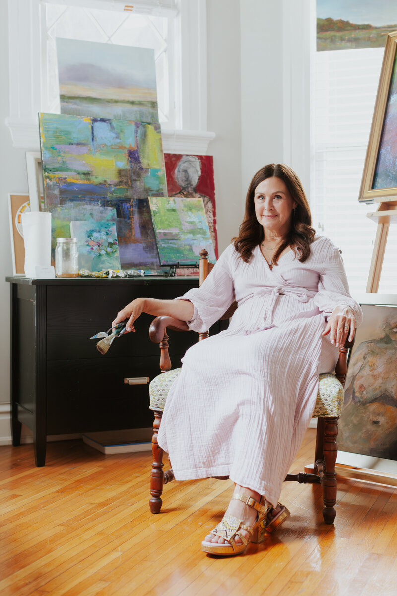 Seated lifestyle headshot of artist Lesley Downey holding paint brushes surrounded by paintings at StudioWorks Gallery in Dilworth Charlotte