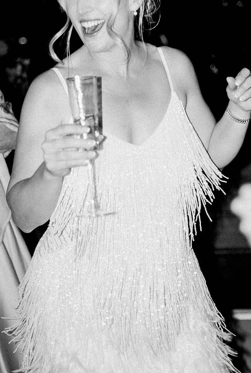 Black and white candid of wedding CA bride dancing with champagne in a glittering fringe dress, captured in luxury documentary wedding photography style.