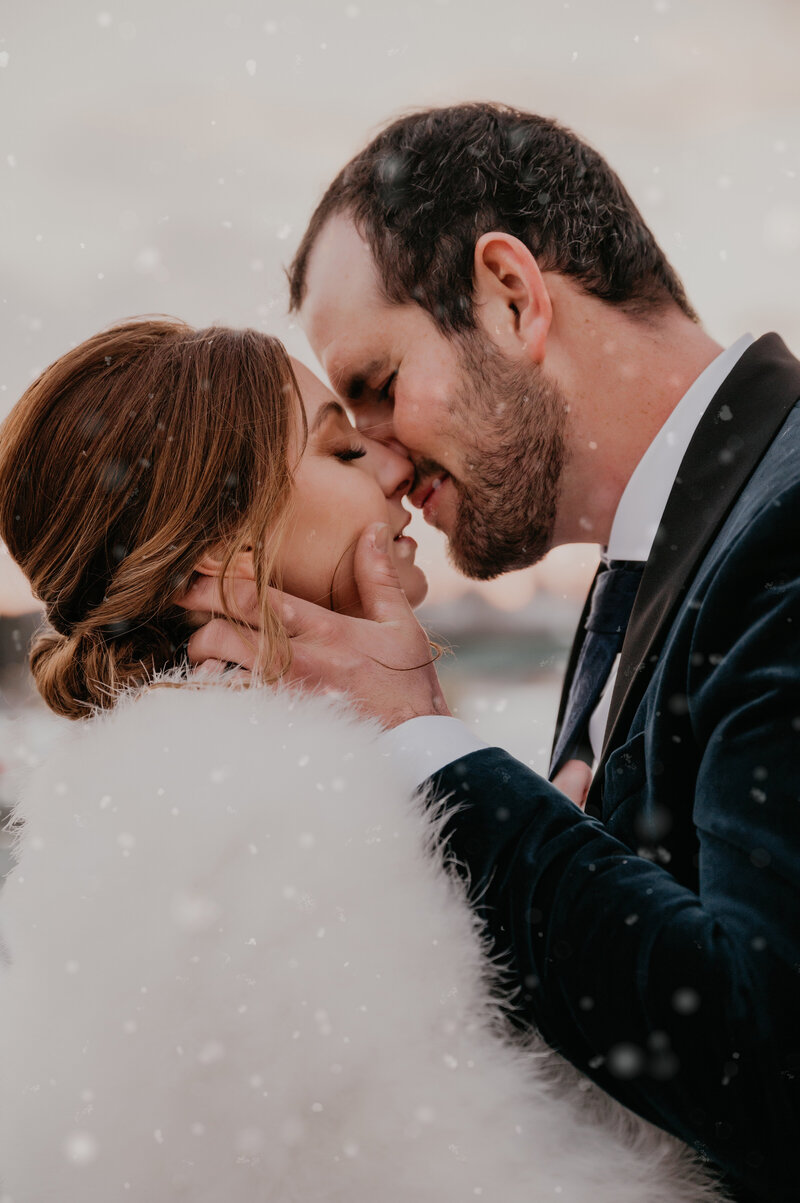 Bride and groom share an intimate kiss at their Elora Mill winter wedding. The groom is holding the bride's cheeks, their noses are touching, and lips parted. The bride is wearing a faux fur stole and the groom in a navy velvet suit. Captured by top KW wedding photographer Ashlee Ellison.