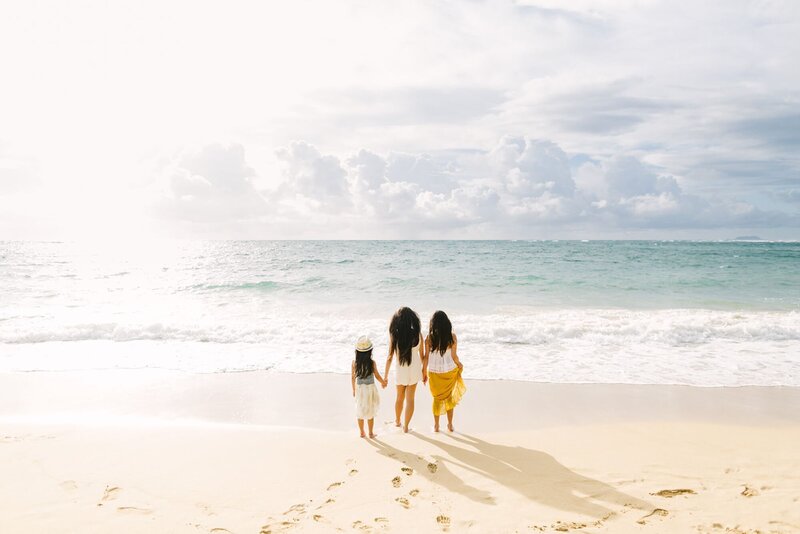 Three young girls hold hands on the beach, looking out over the blue water of North shore.